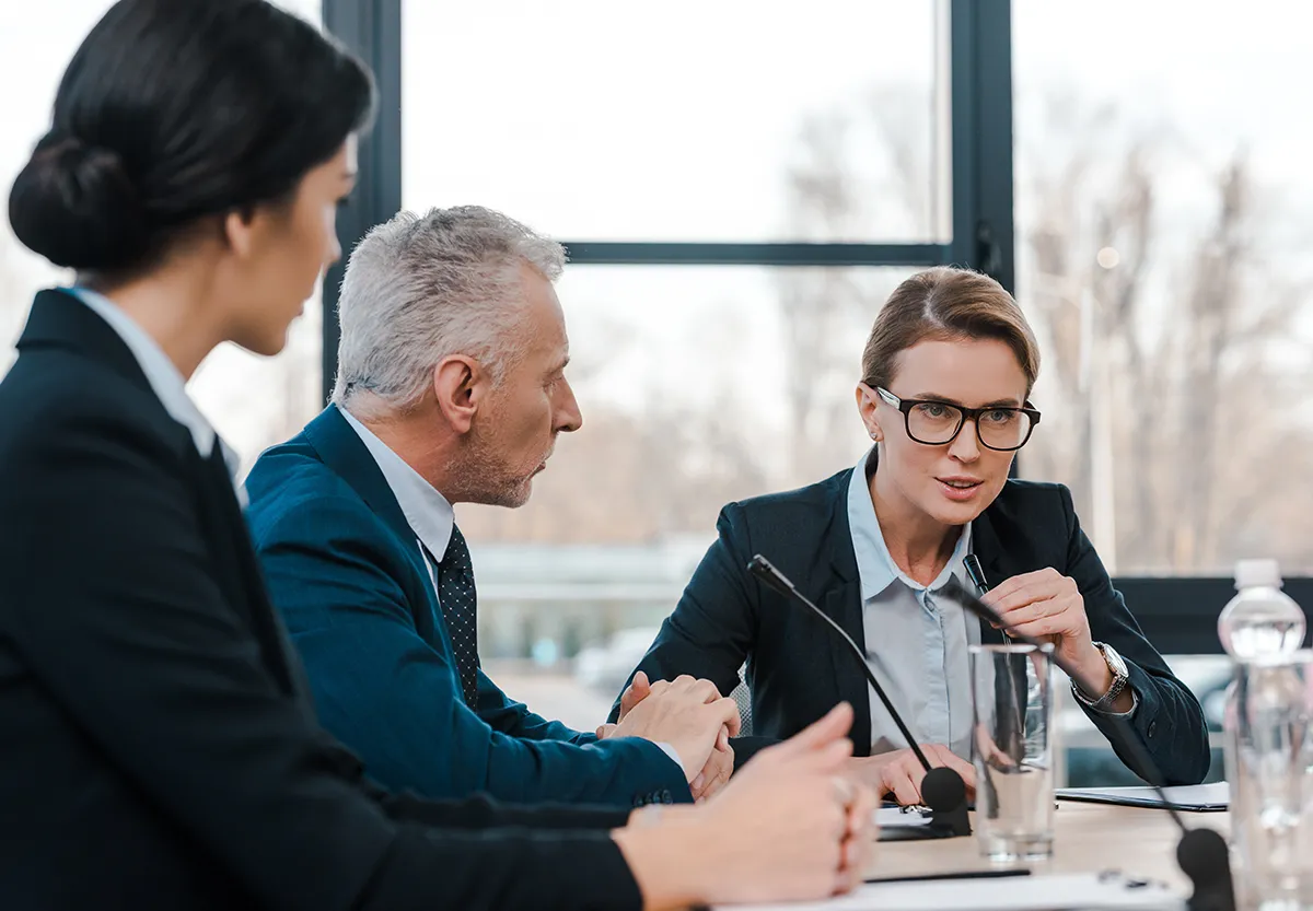 Tre persone in abiti formali in una riunione concentrata attorno a un tavolo di conferenza.